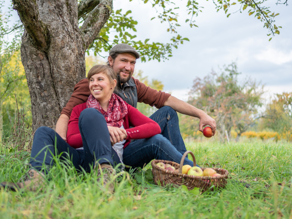 KI generiert: Ein Paar sitzt entspannt unter einem Baum auf einer Wiese, umgeben von einem Korb voller Äpfel. Sie wirken glücklich und genießen offensichtlich einen Moment in der Natur.