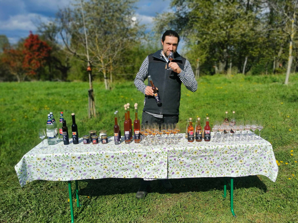 KI generiert: Ein Mann steht im Freien vor einem mit verschiedenen Flaschen und Gläsern gedeckten Tisch, während er an einem Glas riecht. Die Szene wirkt wie eine Verkostung oder Präsentation von Getränken in einer grünen, naturbelassenen Umgebung.