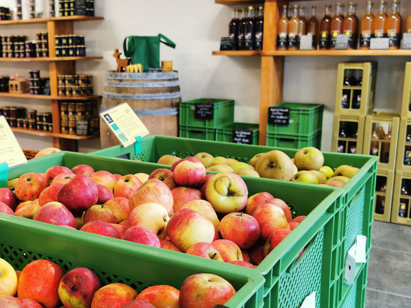 KI generiert: Das Bild zeigt einen Verkaufsraum mit frischen Äpfeln in grünen Kisten im Vordergrund. Im Hintergrund sind Regale mit verschiedenen Flaschen und Lebensmitteln zu sehen.