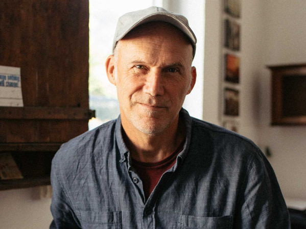 KI generiert: Das Bild zeigt einen Mann in einem blauen Hemd und einer grauen Mütze, der in einem Innenraum vor einer Holzwand steht. Der Raum wirkt gemütlich und hell beleuchtet.