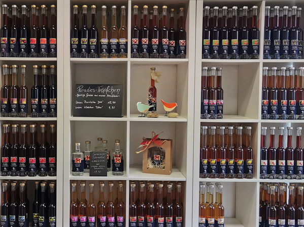 KI generiert: Das Bild zeigt ein Regal mit einer großen Auswahl an farbigen Flaschen, die Liköre oder Spirituosen enthalten. In der Mitte befindet sich eine Tafel mit handgeschriebenen Informationen.